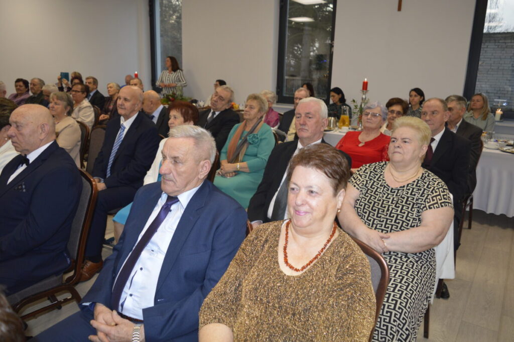 Grupa starszych osób siedzi na przyjęciu w eleganckiej sali. W tle widać duże okna i jasne ściany. Na stołach ustawione są świece i dekoracje.