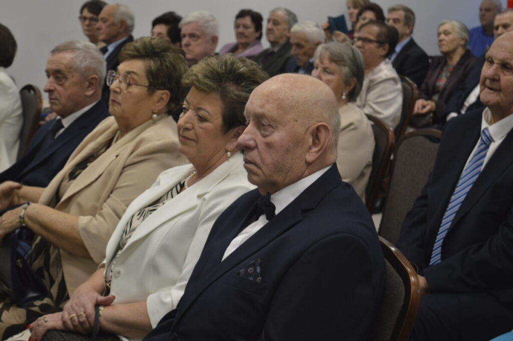 Grupa elegancko ubranych starszych osób siedzi w sali. Ciemne garnitury, jasne garsonki, białe ściany w tle. Uczestnicy skupieni na wydarzeniu.