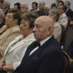 Grupa elegancko ubranych starszych osób siedzi w sali. Ciemne garnitury, jasne garsonki, białe ściany w tle. Uczestnicy skupieni na wydarzeniu.