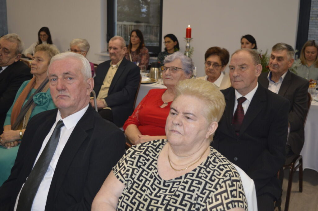 Grupa starszych osób siedzi przy stole w eleganckiej sali. Dominują stonowane kolory, ubrania w odcieniach czerni i szarości. W tle znajduje się czerwona świeca na stole.