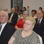 Grupa starszych osób siedzi przy stole w eleganckiej sali. Dominują stonowane kolory, ubrania w odcieniach czerni i szarości. W tle znajduje się czerwona świeca na stole.