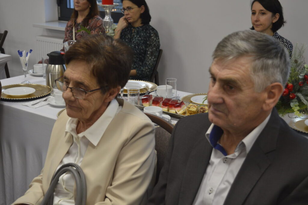 Starsza para siedzi przy stole z białym obrusem, na którym są talerze i desery. W tle kilka osób i dekoracja świąteczna. Atmosfera spotkania towarzyskiego.