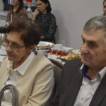 Starsza para siedzi przy stole z białym obrusem, na którym są talerze i desery. W tle kilka osób i dekoracja świąteczna. Atmosfera spotkania towarzyskiego.