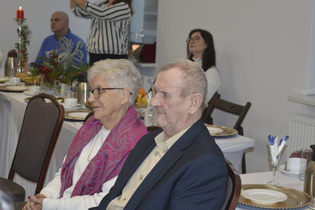 Para starszych ludzi siedzi przy stole na przyjęciu; kobieta ma różowy szal, mężczyzna ciemną marynarkę. Na stole białe obrusy, zastawa i świeczka. W tle siedzą inne osoby.