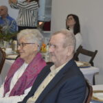 Para starszych ludzi siedzi przy stole na przyjęciu; kobieta ma różowy szal, mężczyzna ciemną marynarkę. Na stole białe obrusy, zastawa i świeczka. W tle siedzą inne osoby.