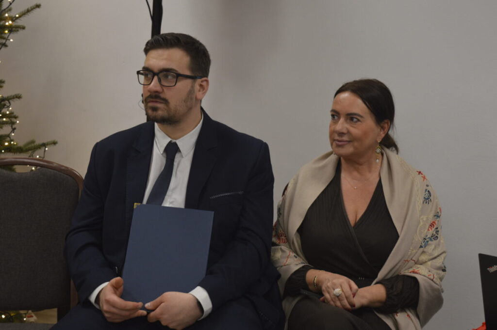 Para siedzi na ciemnoszarych krzesłach w jasnym pomieszczeniu. Obok choinka z lampkami. Kobieta i mężczyzna w eleganckich strojach.