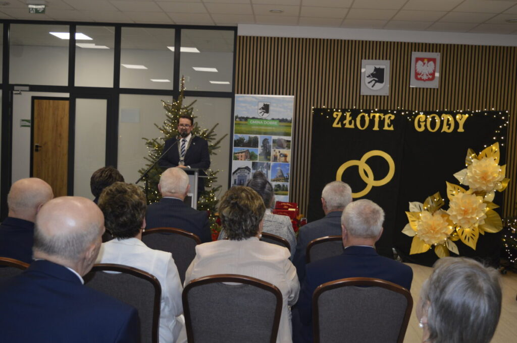 Sala z dekoracją w kolorach złotym i czarnym z napisem ZŁOTE GODY. Mężczyzna przemawia przy mikrofonie. W tle choinka i plakaty.