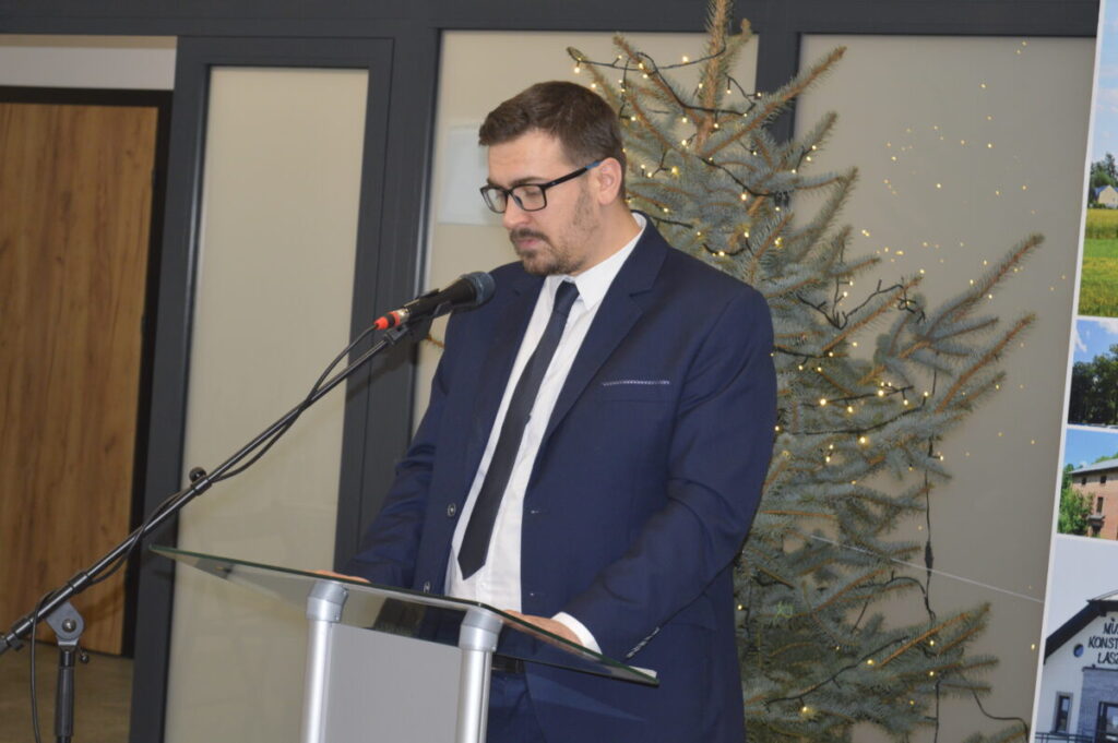 Mężczyzna w granatowym garniturze przemawia przy mikrofonie. W tle widoczna jest ozdobiona choinka. Tło jasne z panelami.