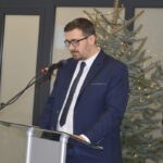 Mężczyzna w granatowym garniturze przemawia przy mikrofonie. W tle widoczna jest ozdobiona choinka. Tło jasne z panelami.