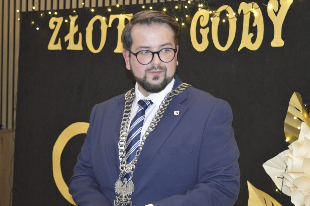 Mężczyzna w granatowym garniturze, nosi łańcuch z emblematem. Tło czarne z napisem Złote Gody, ozdobione światłami. Po prawej papierowe kwiaty.