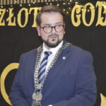 Mężczyzna w granatowym garniturze, nosi łańcuch z emblematem. Tło czarne z napisem Złote Gody, ozdobione światłami. Po prawej papierowe kwiaty.