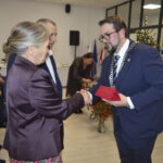 Na zdjęciu kobieta w fioletowej marynarce odbiera nagrodę od mężczyzny w garniturze. Tło jasne, z choinką i plakatem. Widownia siedzi z prawej strony.