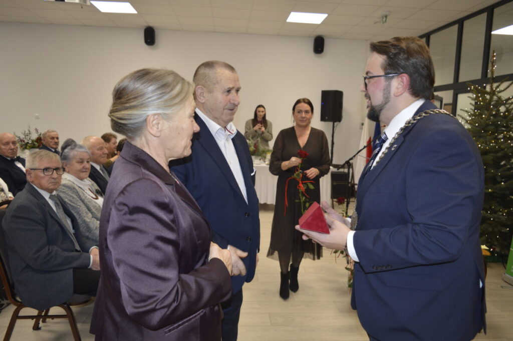 Sala z jasnym sufitem, ozdobiona choinką. Mężczyzna w garniturze wręcza pudełko parze starszych osób. W tle siedzi publiczność.