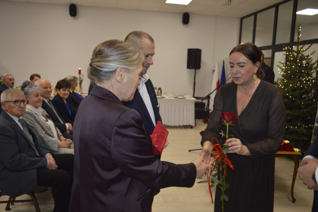 Na zdjęciu widać grupę osób w sali, kobieta wręcza różę. W tle choinka i biało-czerwone tła. Atmosfera oficjalna.