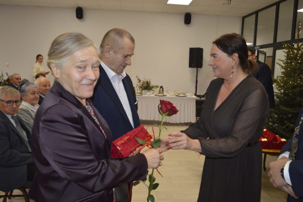 Na zdjęciu dwie kobiety i mężczyzna wymieniają róże w eleganckim wnętrzu. Kobieta po prawej trzyma czerwoną różę. Tło jest jasne z choinką i stołem.