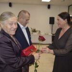 Na zdjęciu dwie kobiety i mężczyzna wymieniają róże w eleganckim wnętrzu. Kobieta po prawej trzyma czerwoną różę. Tło jest jasne z choinką i stołem.