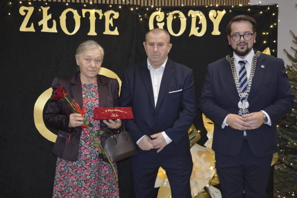 Na zdjęciu troje osób w eleganckich strojach. Starsza kobieta trzyma czerwoną różę, obok mężczyzna i urzędnik w łańcuchu. Tło czarne z napisem ZŁOTE GODY i światełkami.