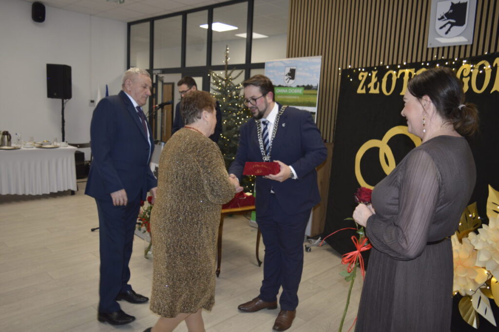 Ludzie w eleganckich strojach uczestniczą w uroczystości wewnątrz. W tle złote obrączki, choinka i dekoracje. Kobieta otrzymuje upominek.