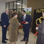 Ludzie w eleganckich strojach uczestniczą w uroczystości wewnątrz. W tle złote obrączki, choinka i dekoracje. Kobieta otrzymuje upominek.