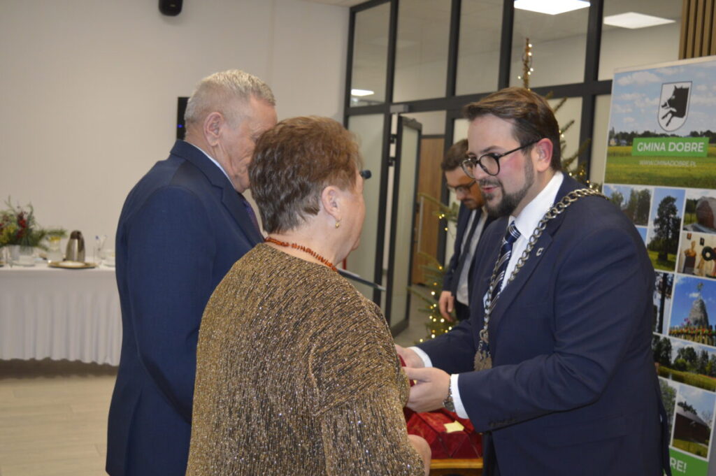 Grupa osób podczas ceremonii w eleganckiej sali, dominują kolory niebieski i złoty. Mężczyzna w garniturze wręcza medal kobiecie. W tle widoczny jest plakat z napisem Gmina Dobre.