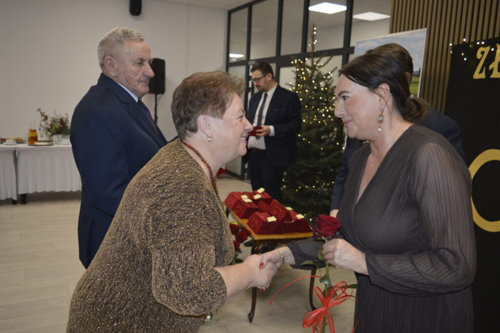 Dwie kobiety podają sobie ręce. Kobieta z prawej trzyma czerwoną różę. W tle choinka i udekorowany stół.