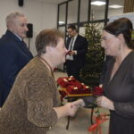 Dwie kobiety podają sobie ręce. Kobieta z prawej trzyma czerwoną różę. W tle choinka i udekorowany stół.