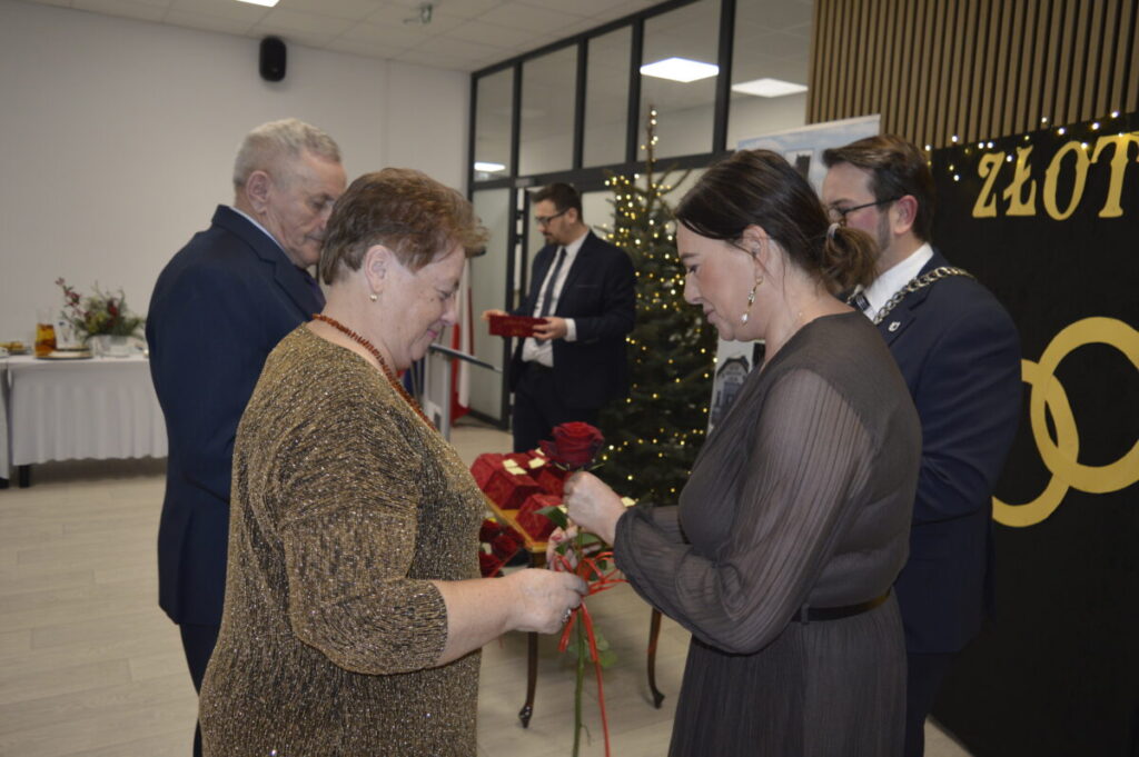 Na zdjęciu grupa ludzi biorących udział w ceremonii. Kobiety trzymają czerwone róże, a w tle widać choinkę i dekoracje. Dominuje elegancki wystrój w złotych i czarnych barwach.