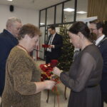 Na zdjęciu grupa ludzi biorących udział w ceremonii. Kobiety trzymają czerwone róże, a w tle widać choinkę i dekoracje. Dominuje elegancki wystrój w złotych i czarnych barwach.