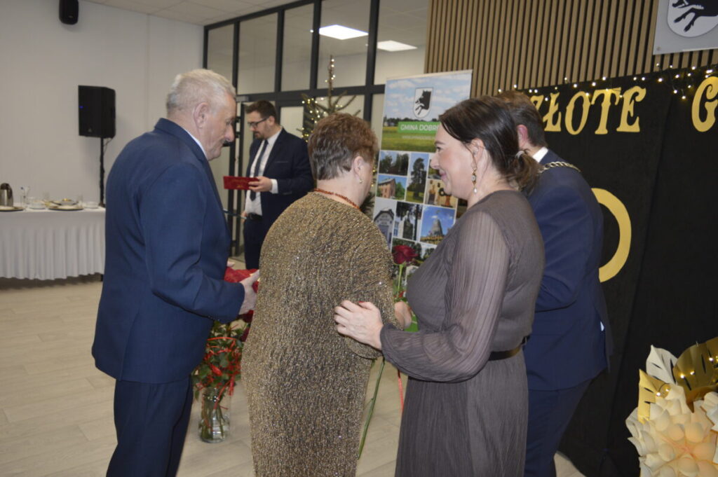 Na zdjęciu są cztery osoby w eleganckich strojach podczas oficjalnej ceremonii. Tło stanowią złote napisy i dekoracje. Kolory dominujące to ciemne odcienie ubrań oraz jasne dekoracje.