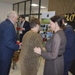 Na zdjęciu są cztery osoby w eleganckich strojach podczas oficjalnej ceremonii. Tło stanowią złote napisy i dekoracje. Kolory dominujące to ciemne odcienie ubrań oraz jasne dekoracje.