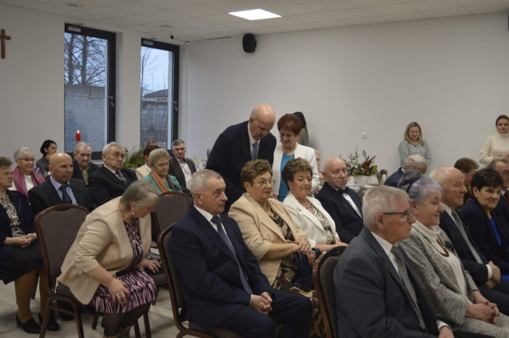 Zdjęcie przedstawia grupę starszych osób siedzących w sali, w jasnym wnętrzu z dużymi oknami. Uczestnicy są elegancko ubrani, tło minimalistyczne. Obok okien widoczna jest dekoracja z kwiatami.