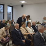 Zdjęcie przedstawia grupę starszych osób siedzących w sali, w jasnym wnętrzu z dużymi oknami. Uczestnicy są elegancko ubrani, tło minimalistyczne. Obok okien widoczna jest dekoracja z kwiatami.