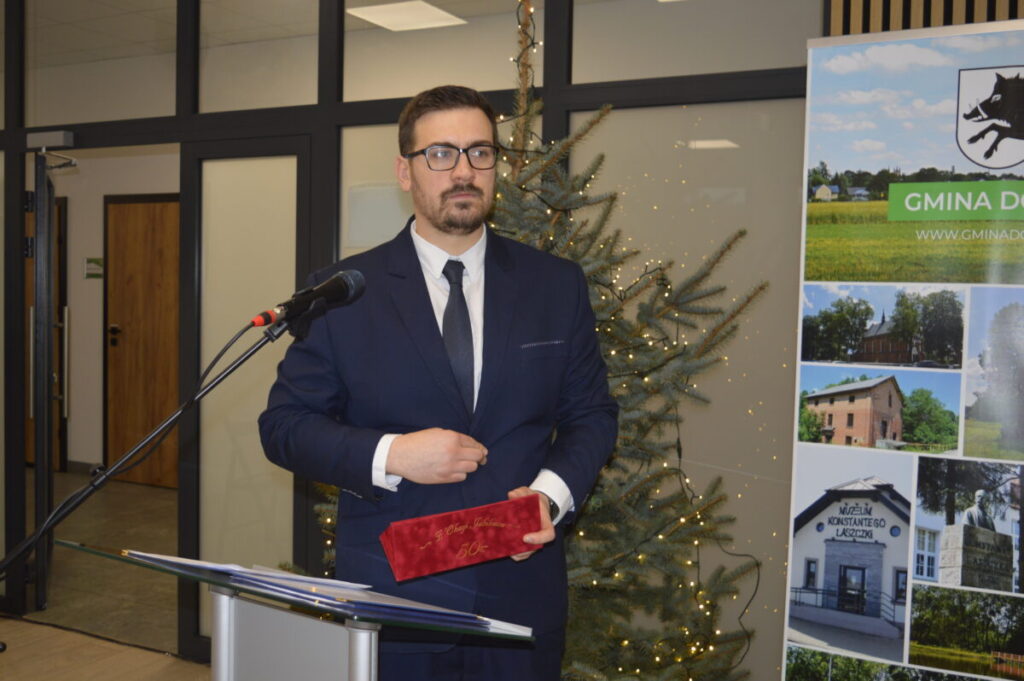 Mężczyzna w garniturze stoi przy mikrofonie z nagrodą w ręce. W tle widać choinkę z lampkami. Obok niego plakat z widokami gminy.