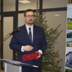 Mężczyzna w garniturze stoi przy mikrofonie z nagrodą w ręce. W tle widać choinkę z lampkami. Obok niego plakat z widokami gminy.