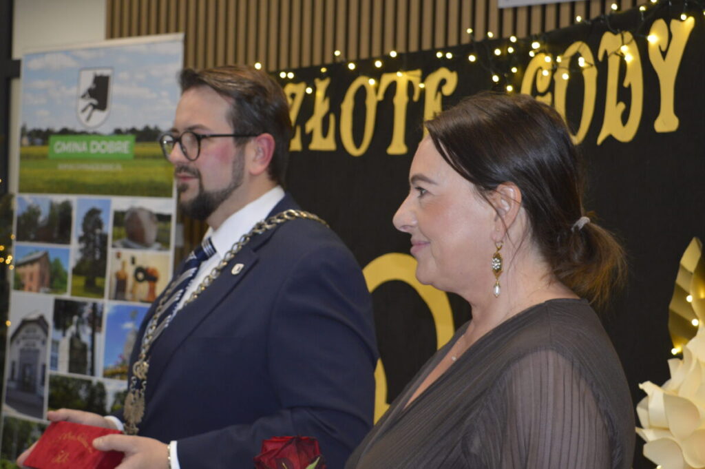 Para stoi przed tłem z napisem Złote Gody, ubrani elegancko, trzymając czerwone róże. Tło jest czarne z żółtymi literami i światełkami. Tablica po lewej zawiera krajobrazy z napisem Gmina Dobre.