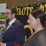 Para stoi przed tłem z napisem Złote Gody, ubrani elegancko, trzymając czerwone róże. Tło jest czarne z żółtymi literami i światełkami. Tablica po lewej zawiera krajobrazy z napisem Gmina Dobre.