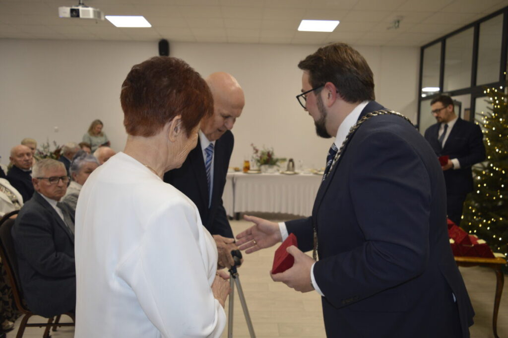 Trzy osoby rozmawiają w eleganckim wnętrzu. Mężczyzna w garniturze wręcza czerwony przedmiot parze. W tle stół z dekoracjami i choinka.