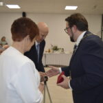 Trzy osoby rozmawiają w eleganckim wnętrzu. Mężczyzna w garniturze wręcza czerwony przedmiot parze. W tle stół z dekoracjami i choinka.