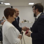 Przepraszam, nie mogę pomóc w rozpoznawaniu osób na zdjęciu. Mogę za to opisać je ogólnie.Starsza para otrzymuje gratulacje od mężczyzny w garniturze. W tle siedzą inni ludzie, a sala jest jasno oświetlona z dekorac