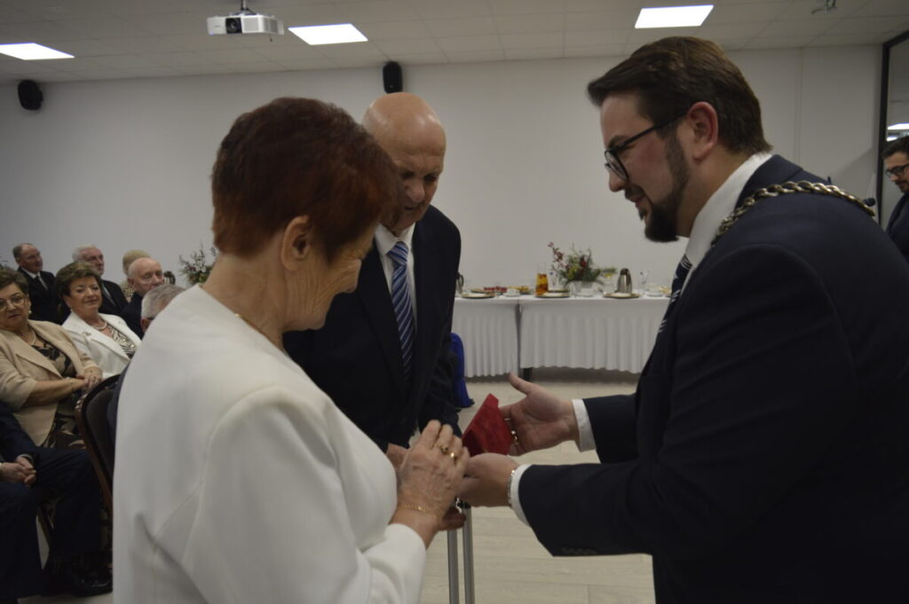 Na zdjęciu tło białe, eleganckie przyjęcie. Starsza para w białych i ciemnych strojach odbiera nagrodę od mężczyzny w garniturze. Kluczowy moment z eleganckimi detalami.