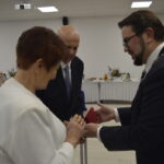 Na zdjęciu tło białe, eleganckie przyjęcie. Starsza para w białych i ciemnych strojach odbiera nagrodę od mężczyzny w garniturze. Kluczowy moment z eleganckimi detalami.