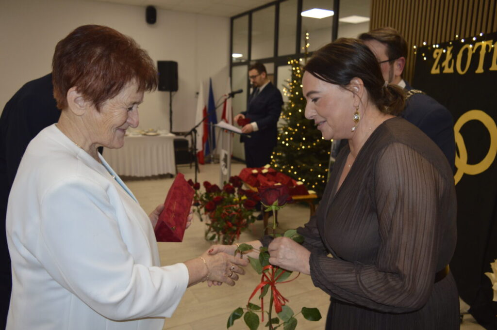 Dwie kobiety wymieniają uściski dłoni, jedna wręcza różę. W tle choinka z lampkami i mężczyzna przy mikrofonie.