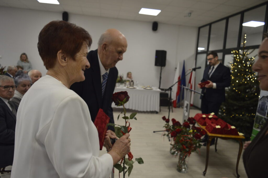Starsza para odbiera róże na uroczystości w jasnej sali. Obok widoczny mężczyzna przy mikrofonie. W tle stoi oświetlona choinka.