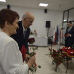 Starsza para odbiera róże na uroczystości w jasnej sali. Obok widoczny mężczyzna przy mikrofonie. W tle stoi oświetlona choinka.