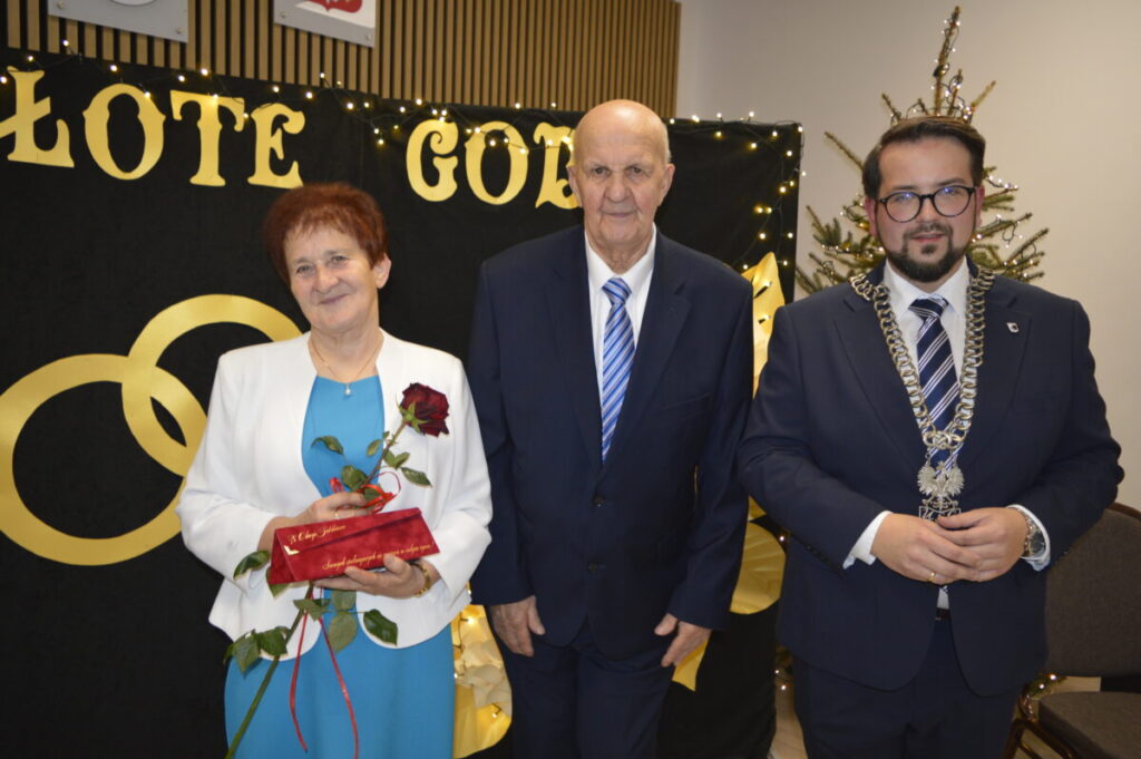 Zdjęcie pokazuje trzy osoby na tle napisu ZŁOTE GODY. Kobieta trzyma różę i prezent. Tło ozdobione światełkami, w rogu widać choinkę.