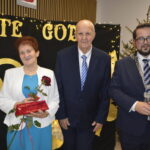 Zdjęcie pokazuje trzy osoby na tle napisu ZŁOTE GODY. Kobieta trzyma różę i prezent. Tło ozdobione światełkami, w rogu widać choinkę.