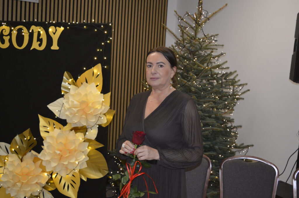 Kobieta w ciemnej sukni trzyma czerwoną różę, stojąc obok choinki i ozdób. Tło czarne z żółtymi literami. Dekoracje świetlne.