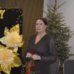 Kobieta w ciemnej sukni trzyma czerwoną różę, stojąc obok choinki i ozdób. Tło czarne z żółtymi literami. Dekoracje świetlne.