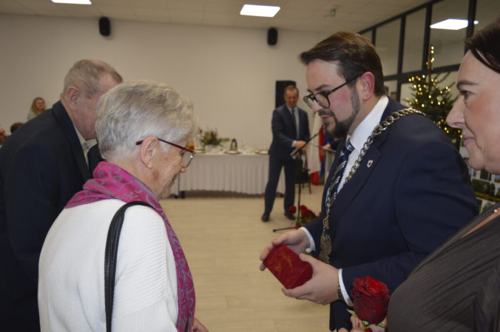 Spotkanie w eleganckim wnętrzu, mężczyzna w garniturze wręcza nagrodę starszej kobiecie w różowym szaliku. W tle choinka.