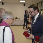 Spotkanie w eleganckim wnętrzu, mężczyzna w garniturze wręcza nagrodę starszej kobiecie w różowym szaliku. W tle choinka.